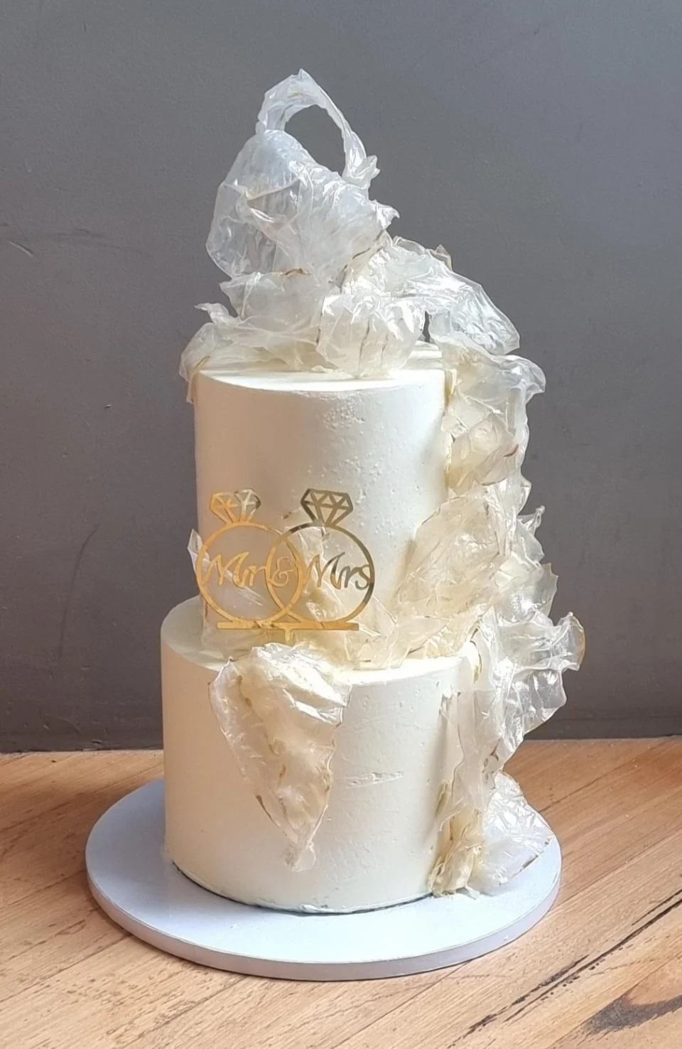 A two-tier white wedding cake decorated with white decorative material and a gold 'Mr & Mrs' topper with ring icons.
