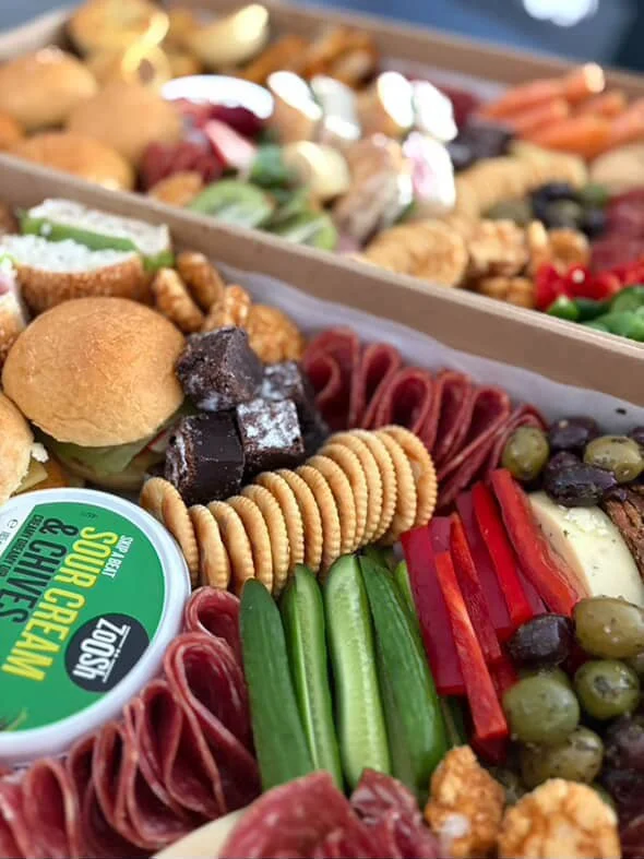 Close-up of a platter with assorted snacks including crackers, green olives, red bell pepper strips, dried meats, grapes, cheese, and a small container of sun-dried tomato spread.