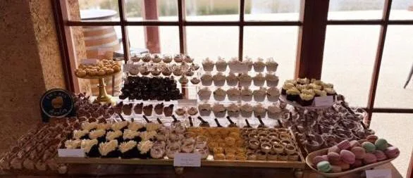Assorted cupcakes and macarons on a dessert table near a window.
