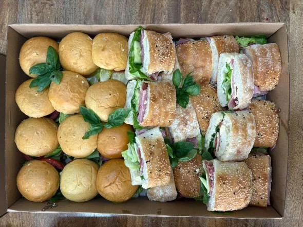 A box containing several mini sandwiches with deli meat, lettuce, and cheese, and an assortment of small, round bread rolls garnished with fresh basil leaves.