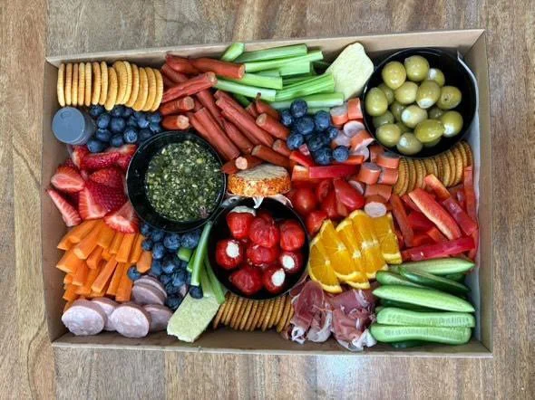 A large tray filled with various fresh vegetables, fruits, crackers, and dips, including celery sticks, blueberries, strawberries, cherry tomatoes, sliced bell peppers, cucumbers, green olives, radishes, and a bowl of green dip.