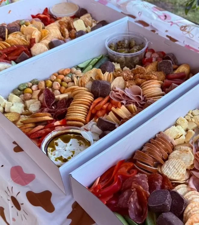 A white tray of assorted snacks including crackers, charcuterie, cheese, grapes, and dips.
