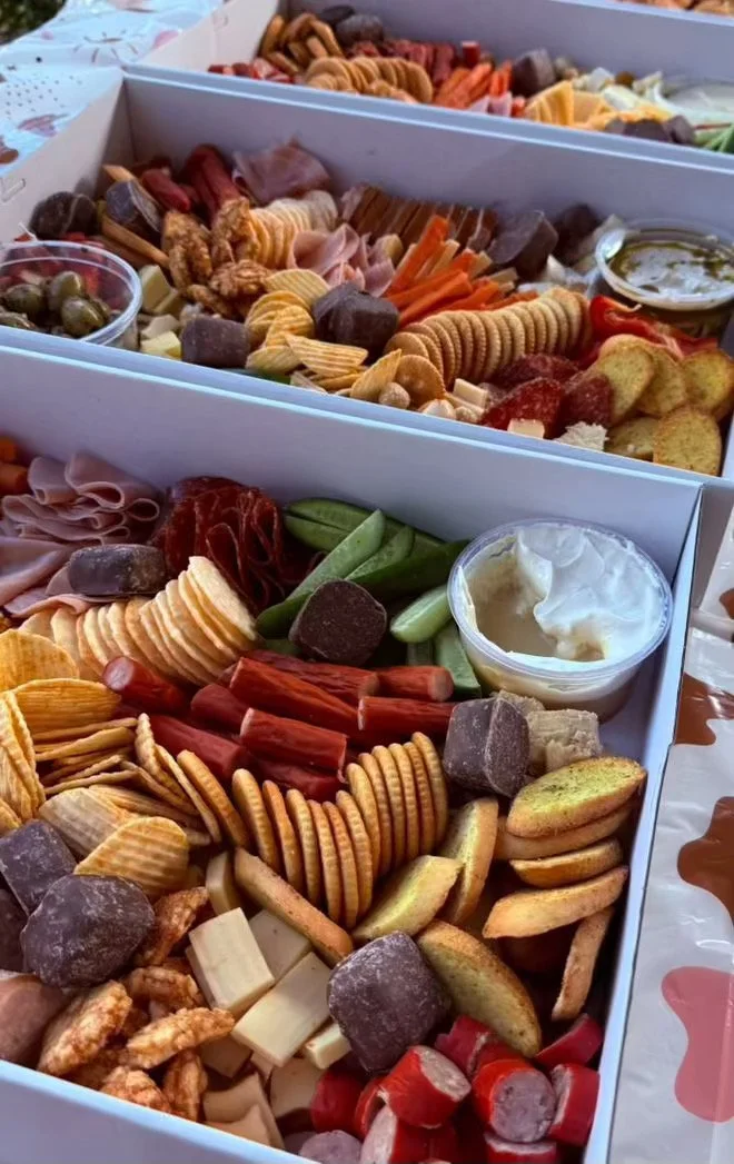 Two white boxes filled with assorted snacks including crackers, pretzels, cheese cubes, sliced meats, cookies, potato chips, vegetables, and dips.