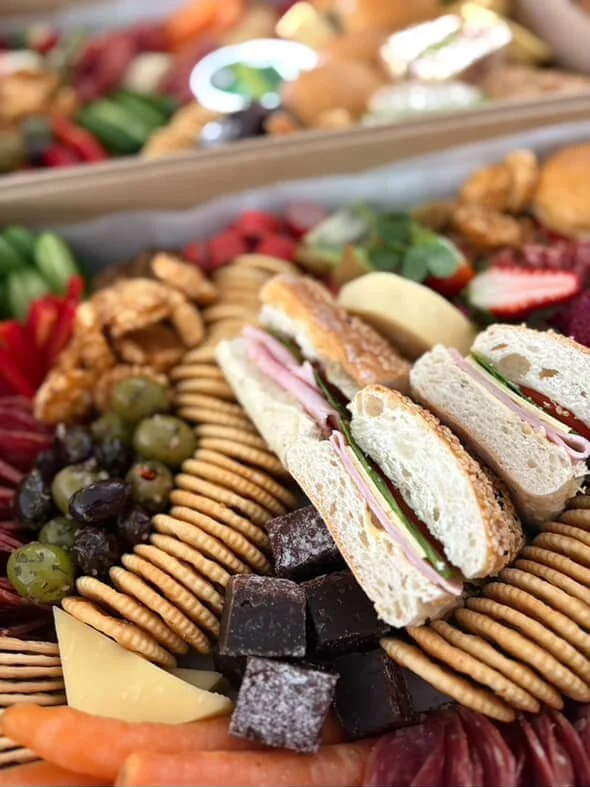 Assorted finger foods on a platter including sandwiches, crackers, green and black olives, cheese, fruit, and candies.