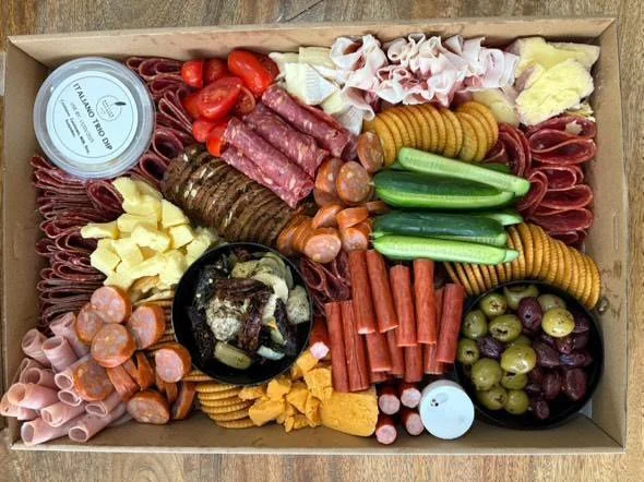 A charcuterie board with various cured meats, cheeses, crackers, cherry tomatoes, green olives, grapes, and dips.