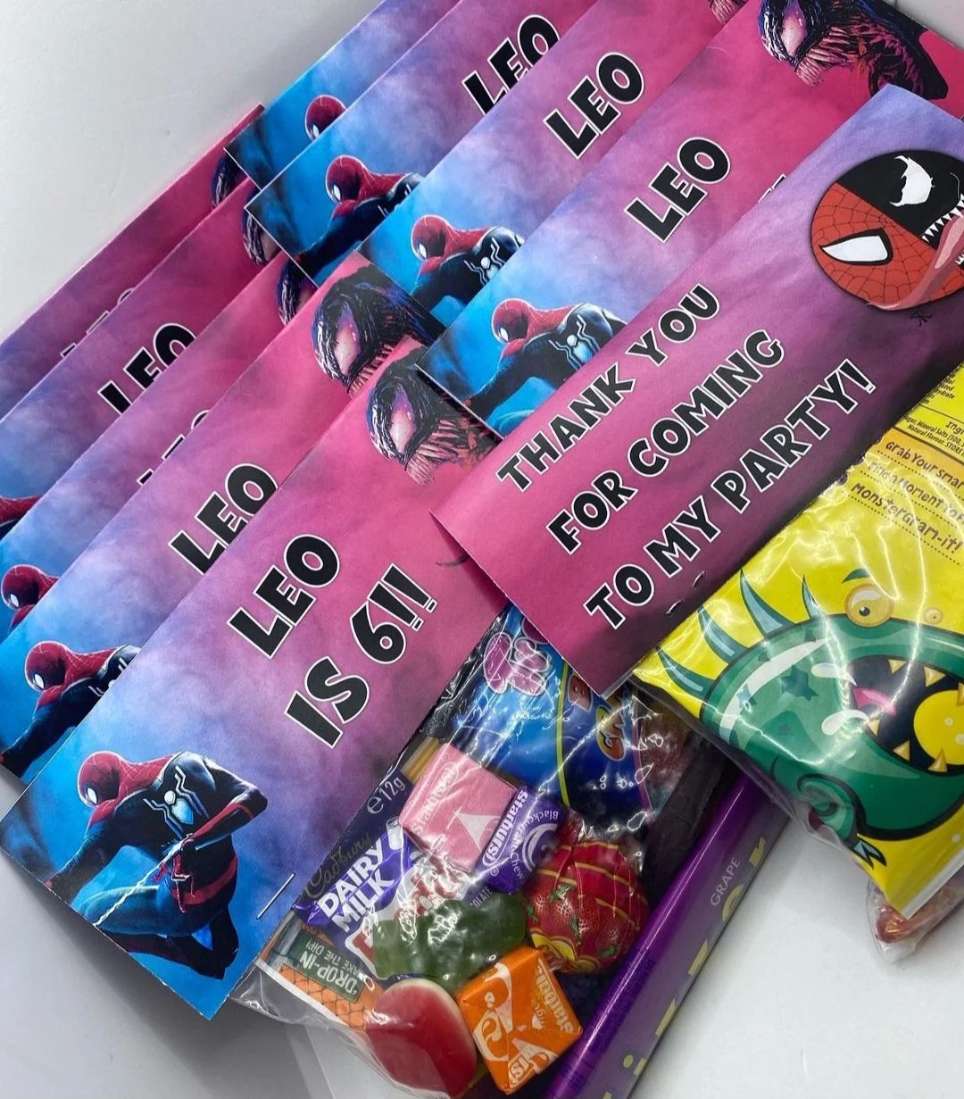 A collection of Spider-Man themed thank you notes with a pink background and pictures of Spider-Man, along with candies and treats, including a yellow bag of gummies and other assorted candies.