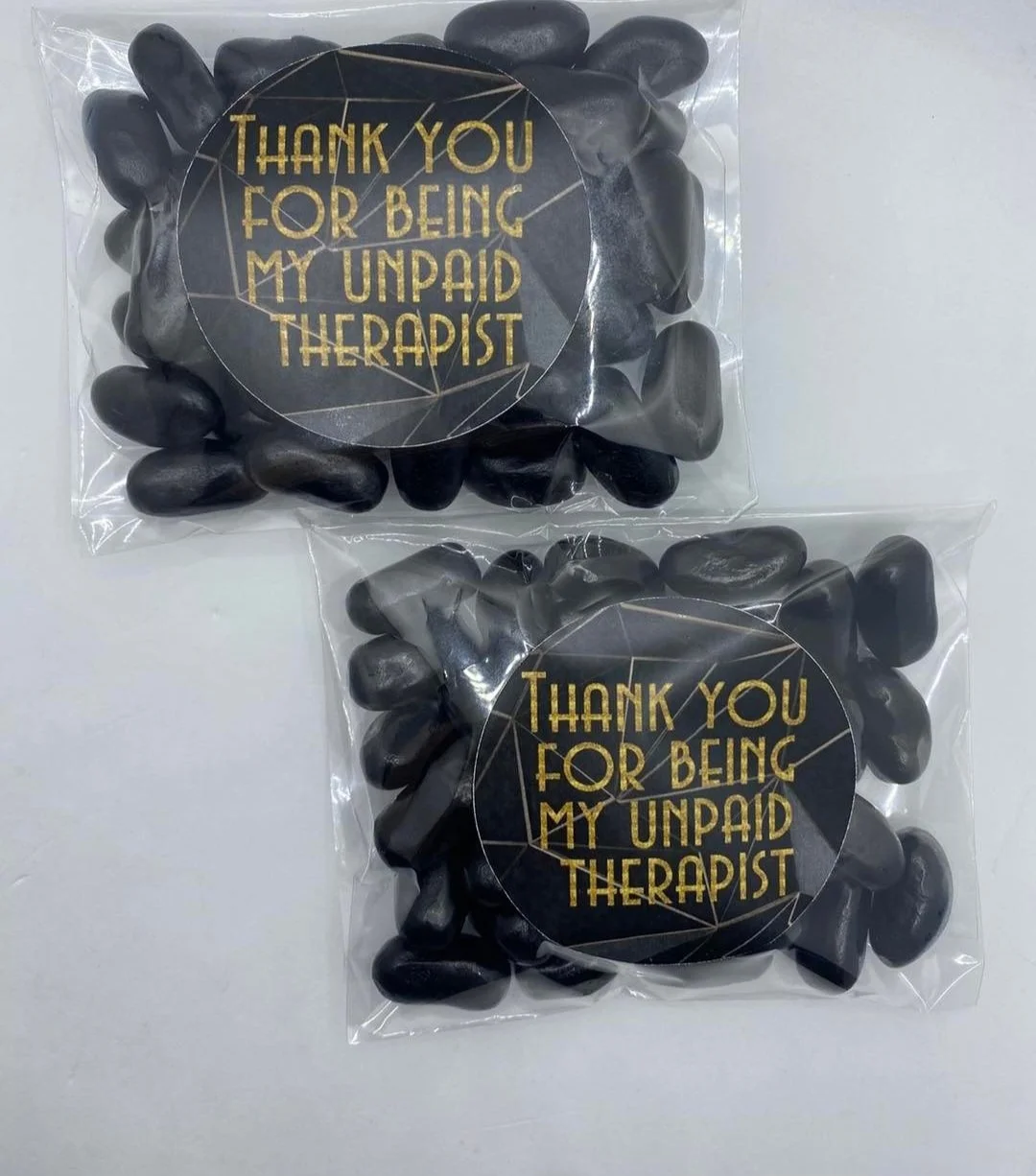 Two clear plastic bags containing black rocks or stones, each with a round black sticker that has gold text reading 'Thank you for being my unpaid therapist'.