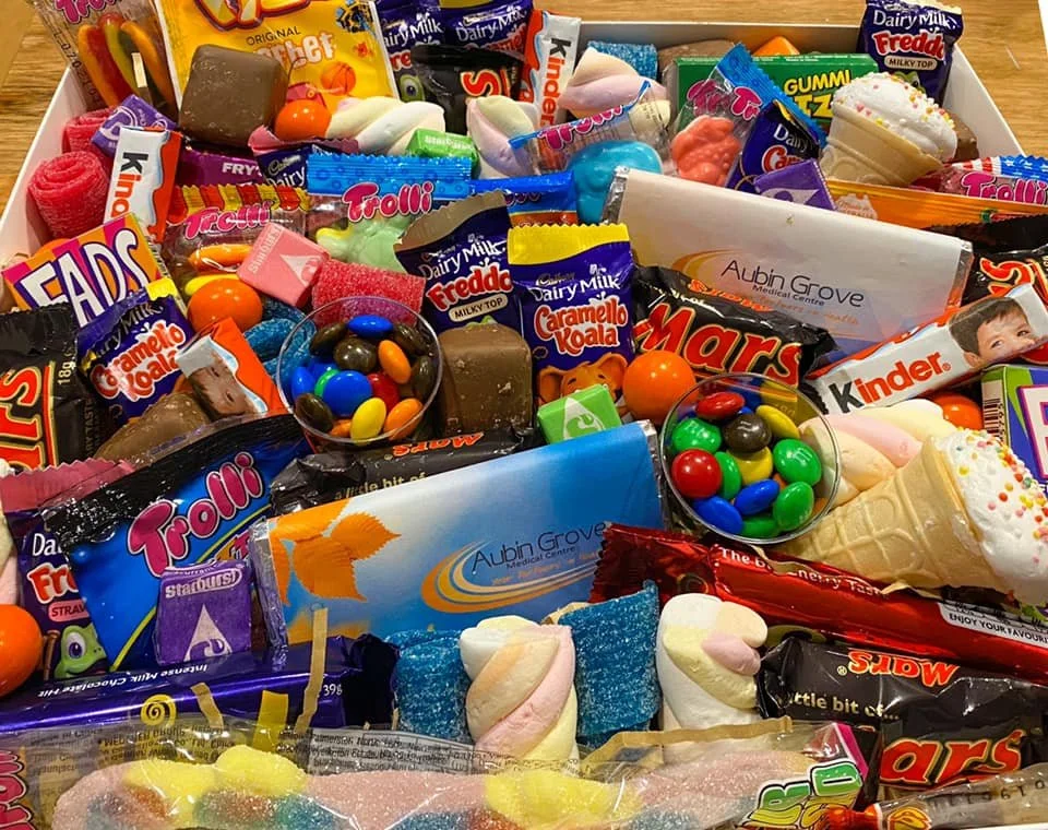 A box filled with assorted candies including chocolate bars, gummy worms, marshmallows, gum, and small round chocolates, along with some ice cream cone-shaped sweets and candies in bright packaging.
