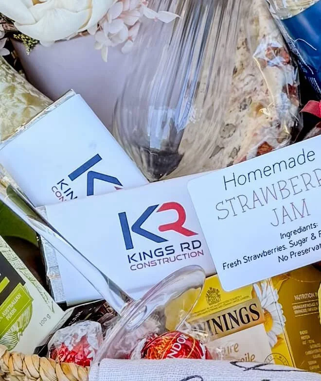 A homemade strawberry jam label with Kings Rd Construction logo, placed among various packaged food items and flowers.