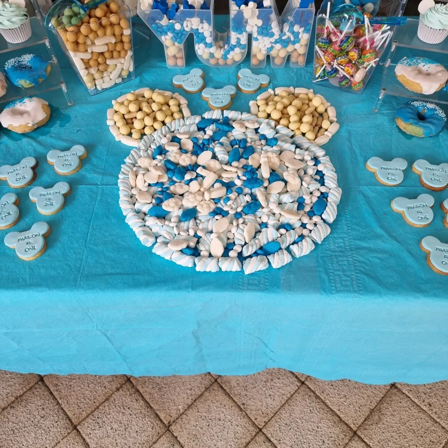 A table decorated with blue and white candies, cookies, and cupcakes for a celebration with a blue tablecloth and a 'MOM' themed candy display.