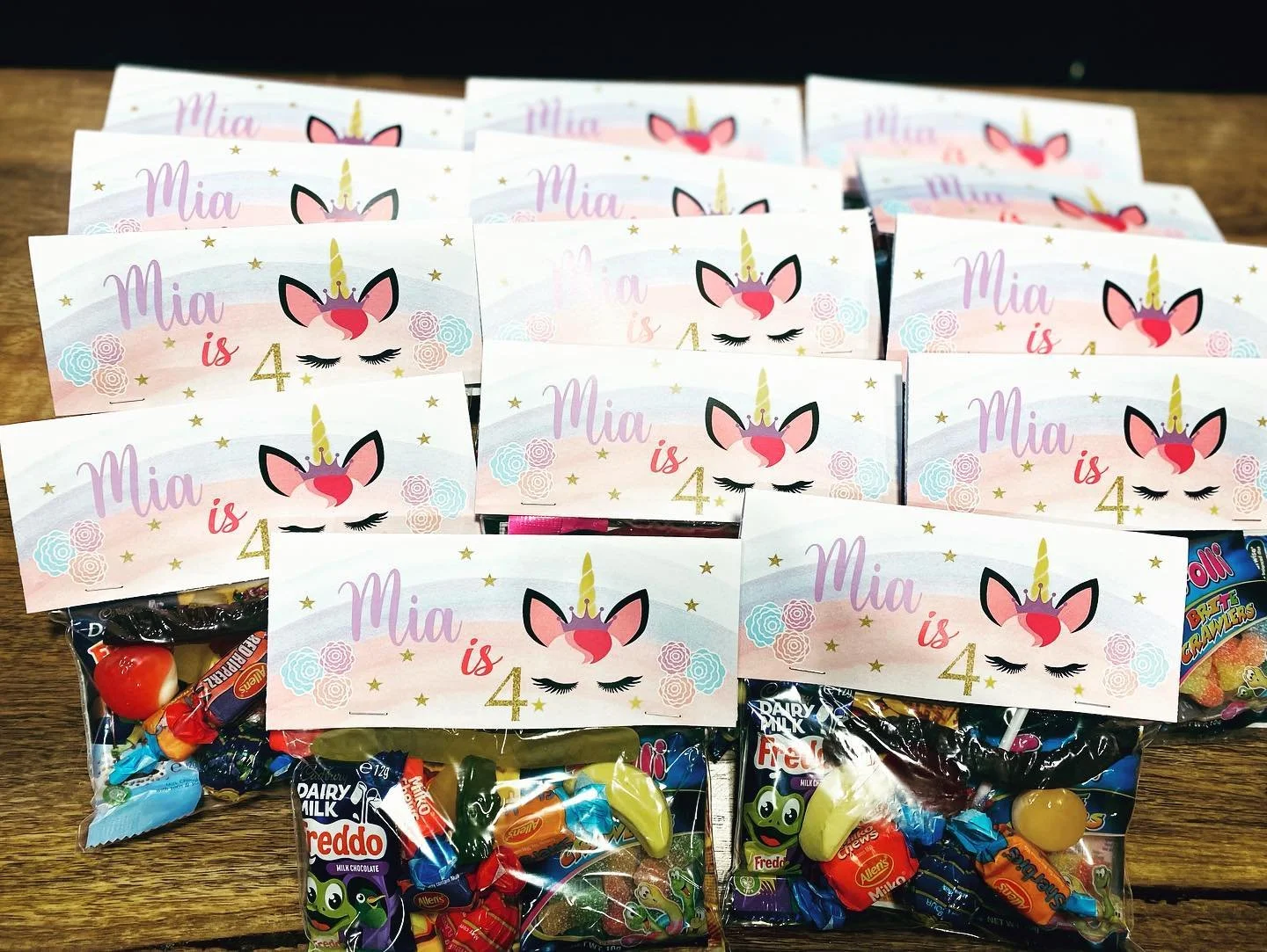 Several birthday cards with unicorn and pastel rainbow design reading 'Mia is 4' arranged on a table, with bags of assorted candies underneath.