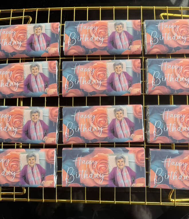 Small rectangular boxes with a birthday greeting, featuring an elderly woman with short gray hair, wearing a pink jacket and a plaid scarf, against a backdrop of large pink and orange roses.