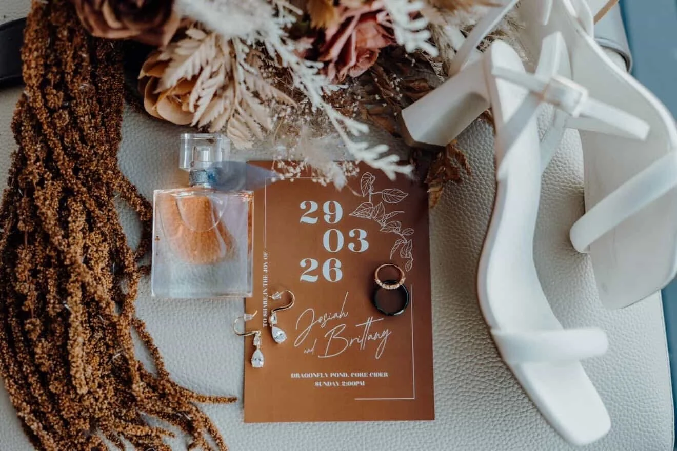 Wedding invitation with earrings, rings, a perfume bottle, and decorative dried flowers on a textured surface.
