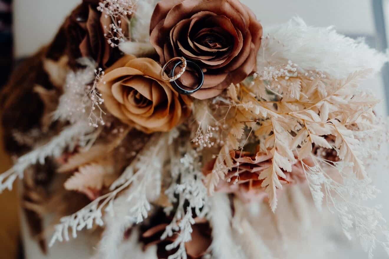Close-up of wedding rings resting on a bouquet of dried roses, leaves, and delicate white flowers.
