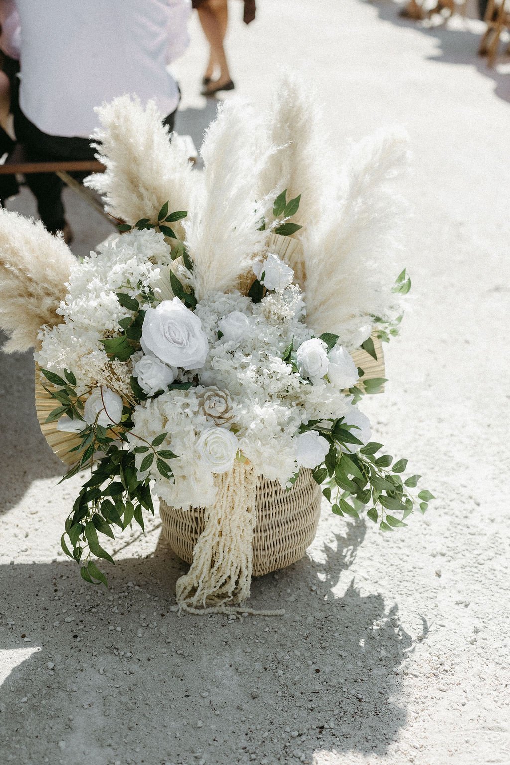 A flower arrangement with white roses, hydrangeas, and pampas grass in a woven basket on a sandy surface.