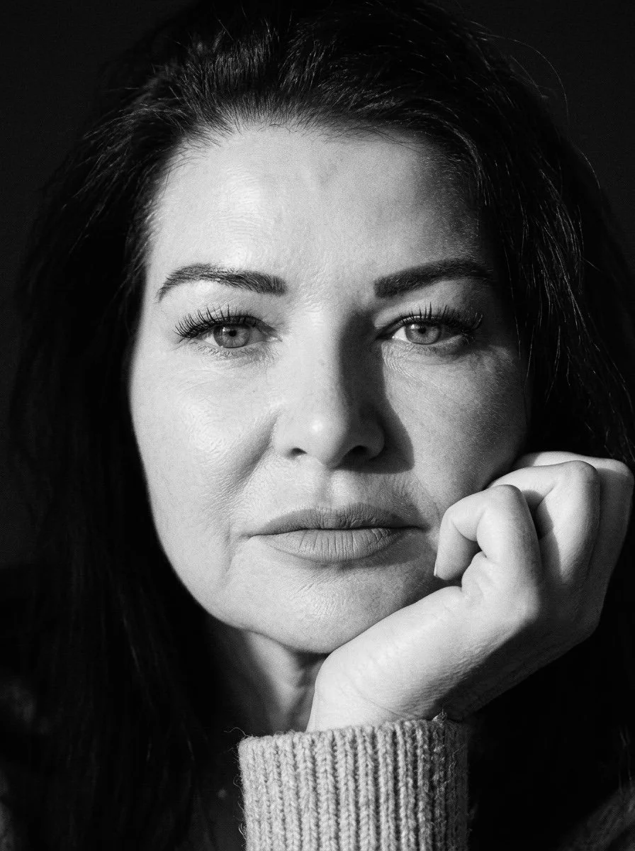 Close-up black and white portrait of a woman with dark hair, gazing directly at the camera, resting her chin on her hand, wearing a sweater.