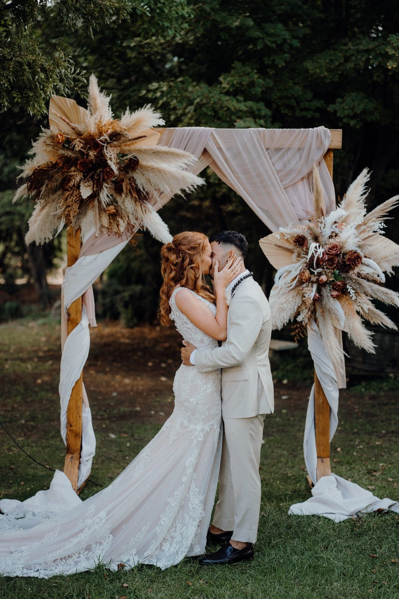 A bride and groom sharing a kiss under a decorated outdoor wedding arch with flowers and drapery, in a lush green natural setting.