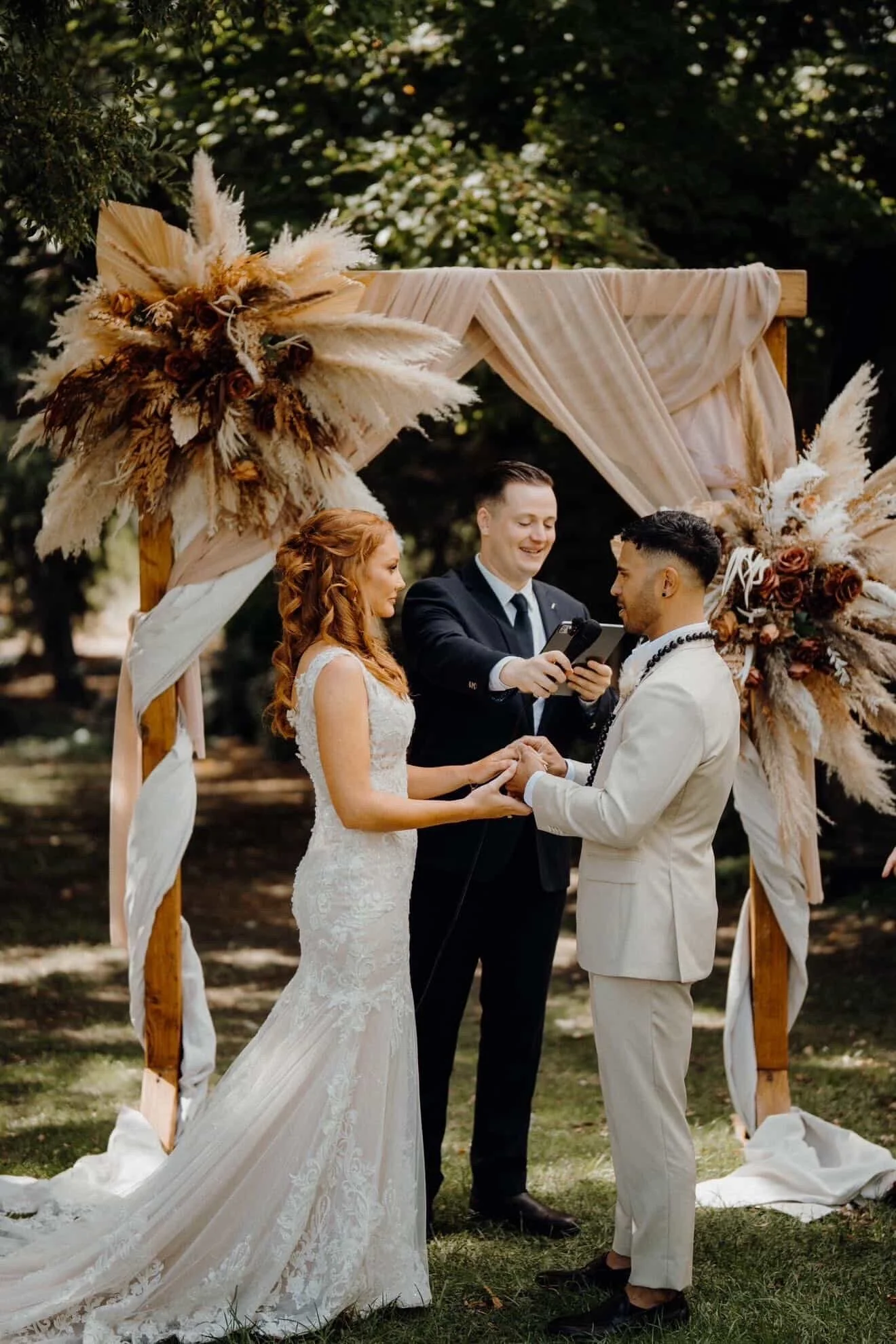 A wedding ceremony outdoors with a bride and groom holding hands, exchanging vows, under an arch decorated with dried flowers and fabric, with an officiant reading to them.