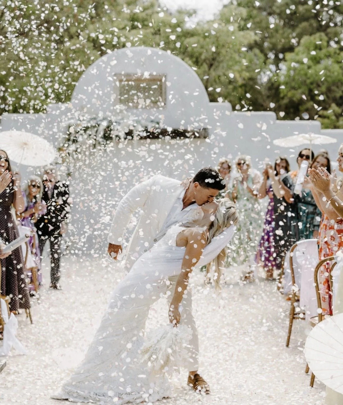 A wedding celebration with a couple kissing amidst falling white flower petals, surrounded by friends and family outdoors with a white building and trees in the background.