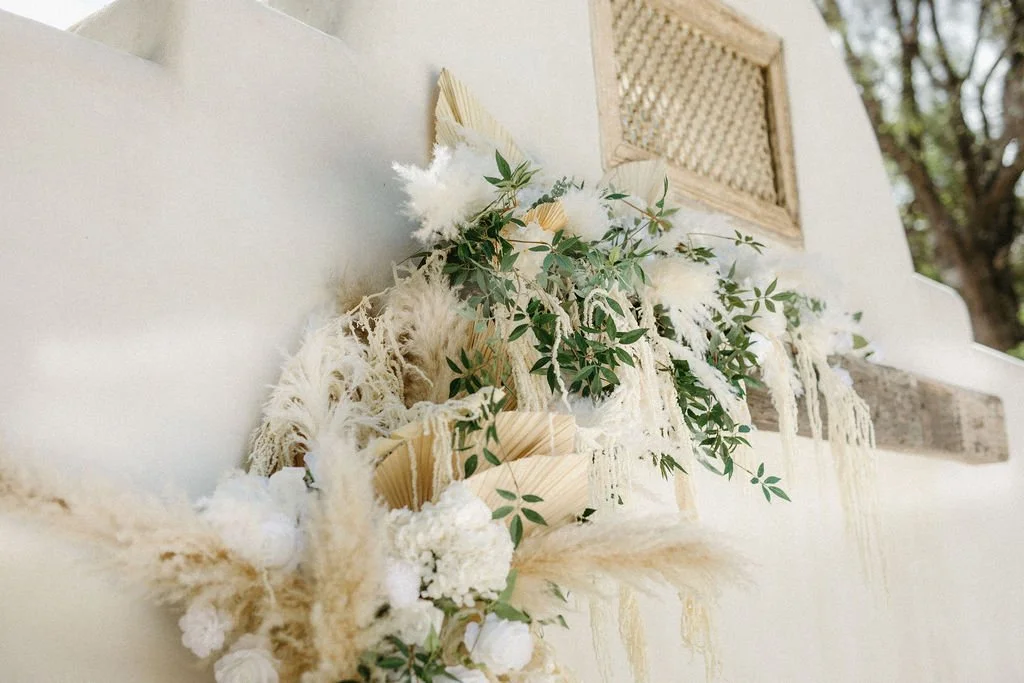 Decorative arrangement of white flowers, greenery, and dried foliage mounted on a white wall with a decorative vent and outdoor trees visible in the background.