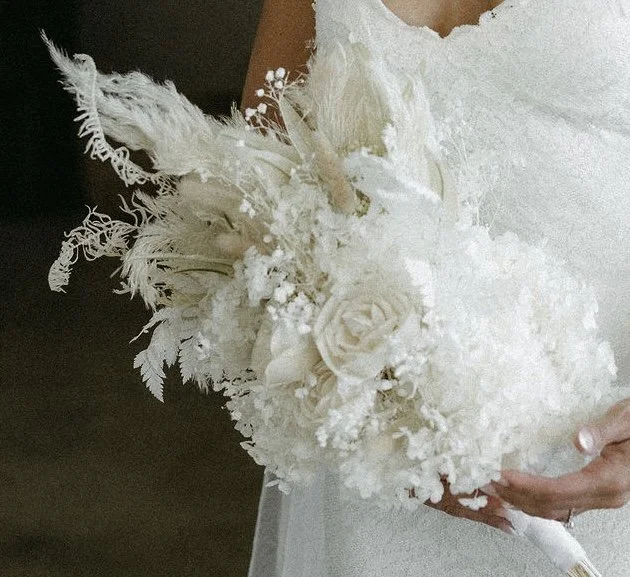 A bride holding a white bouquet of flowers and feathers, dressed in a white gown.
