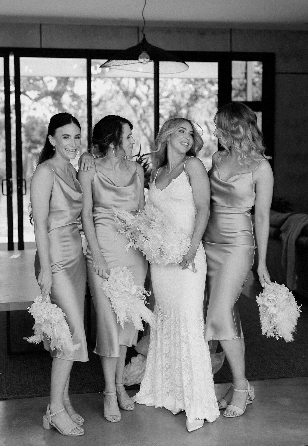 Four women dressed in elegant attire, standing indoors near a large window, smiling and holding bouquets of flowers, possibly at a wedding or special event.