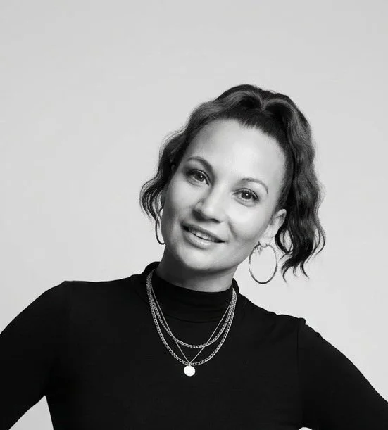Black and white portrait of a woman with wavy hair, wearing hoop earrings, layered necklaces, and a black top with a turtleneck, smiling at the camera.