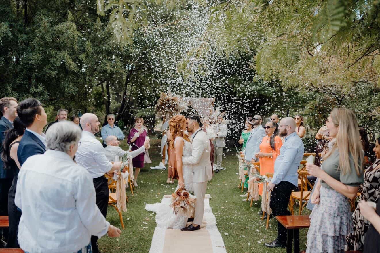 A wedding ceremony outdoors with a bride and groom kissing in the center, surrounded by wedding guests throwing flower petals and celebrating, with trees in the background.