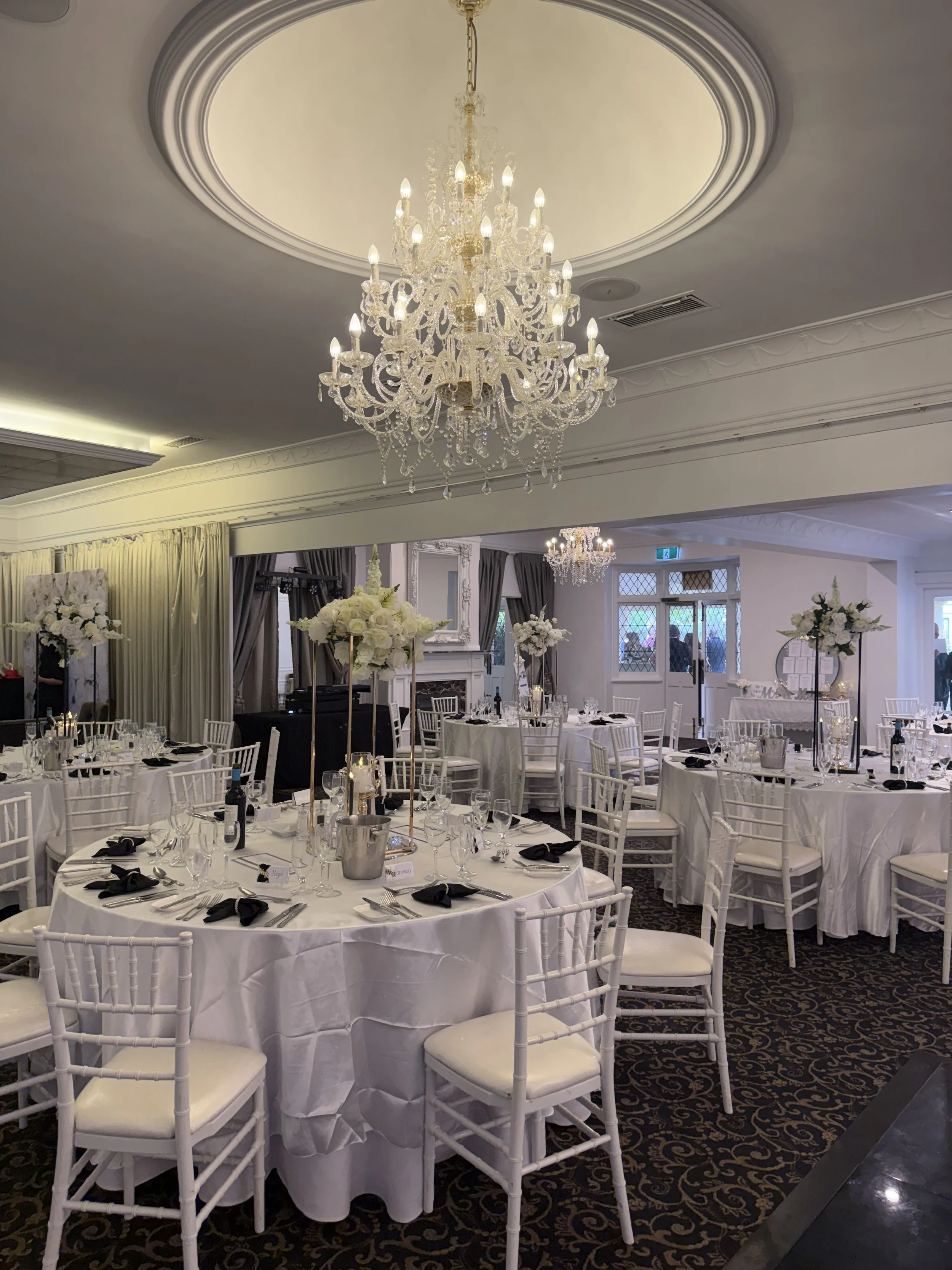 Elegant banquet hall decorated for a special event, featuring a large crystal chandelier hanging from the ceiling, round tables with white tablecloths, black napkins, and floral centerpieces of white flowers in tall vases, surrounded by white chairs with padded seats. The room has white walls, decorated moldings, and windows with curtains.