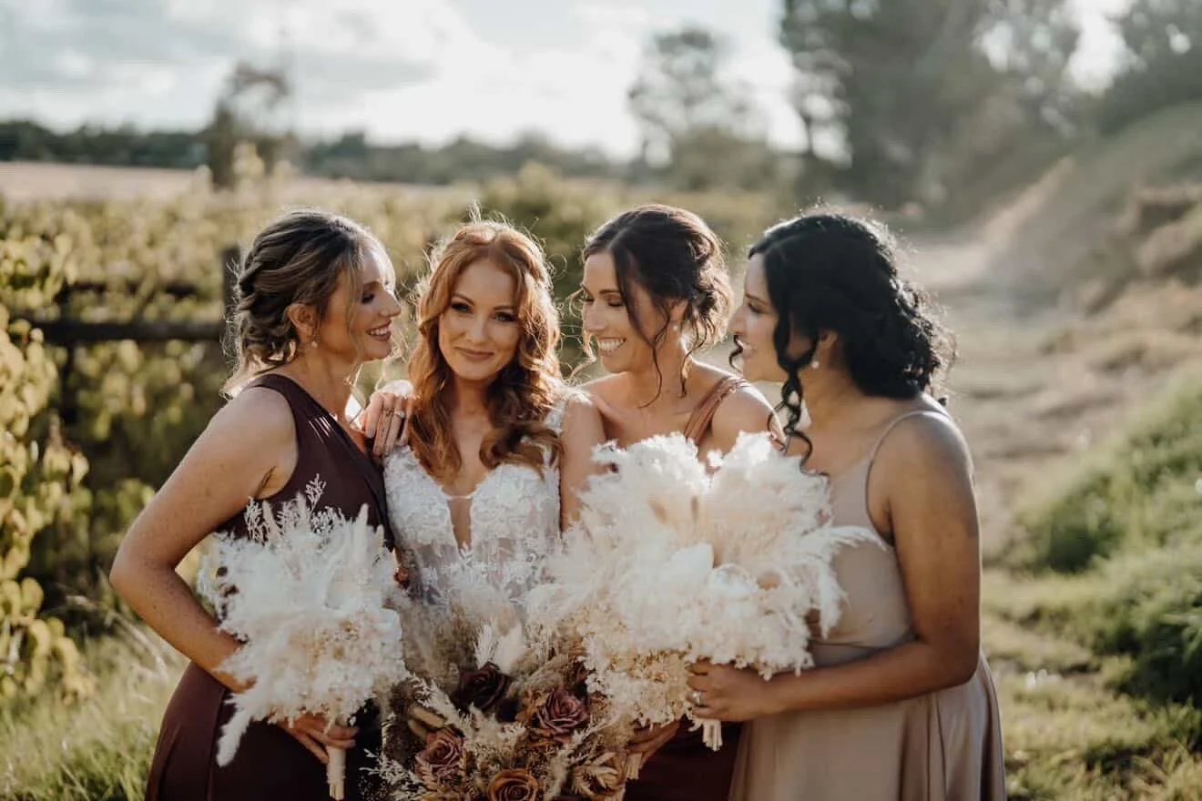 Four women in bridesmaid dresses standing outdoors in a field, smiling and holding bouquets, celebrating a wedding.