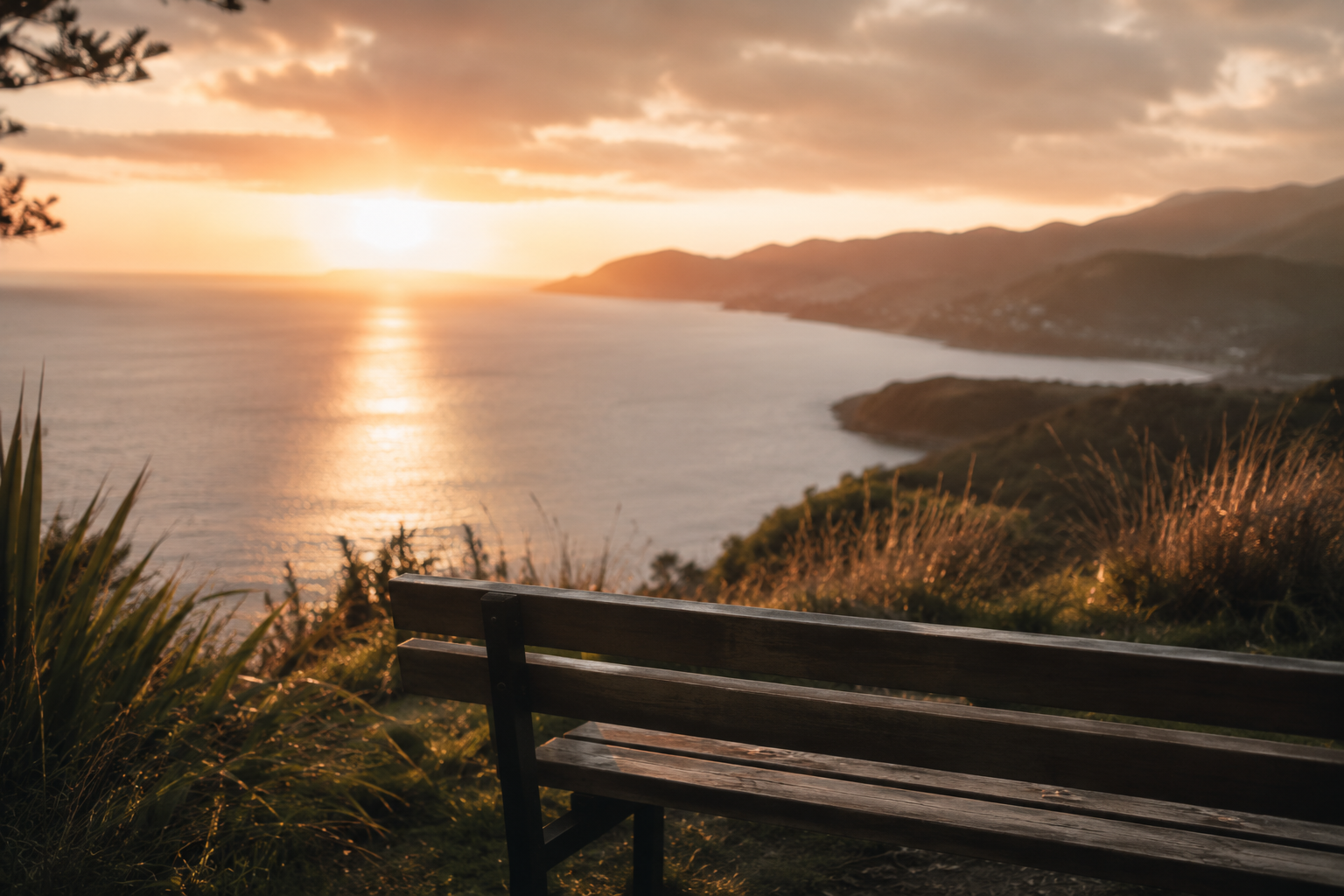 A wooden bench facing a calm body of water during sunset, with hills in the background and clouds in the sky.