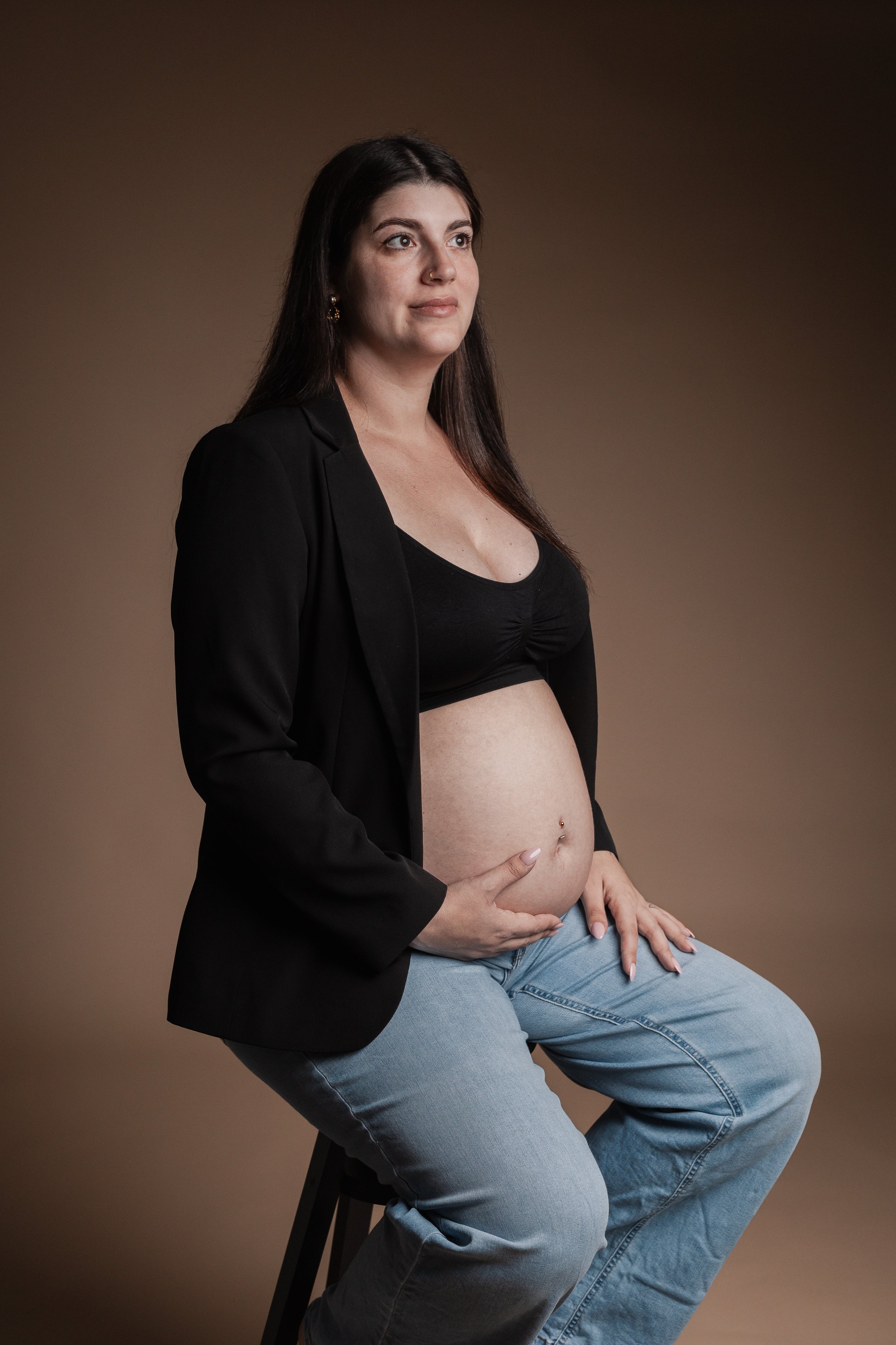 A pregnant woman sitting on a stool, wearing a black crop top and a black blazer, with her hand on her belly, and a neutral expression on her face, against a plain brown background.