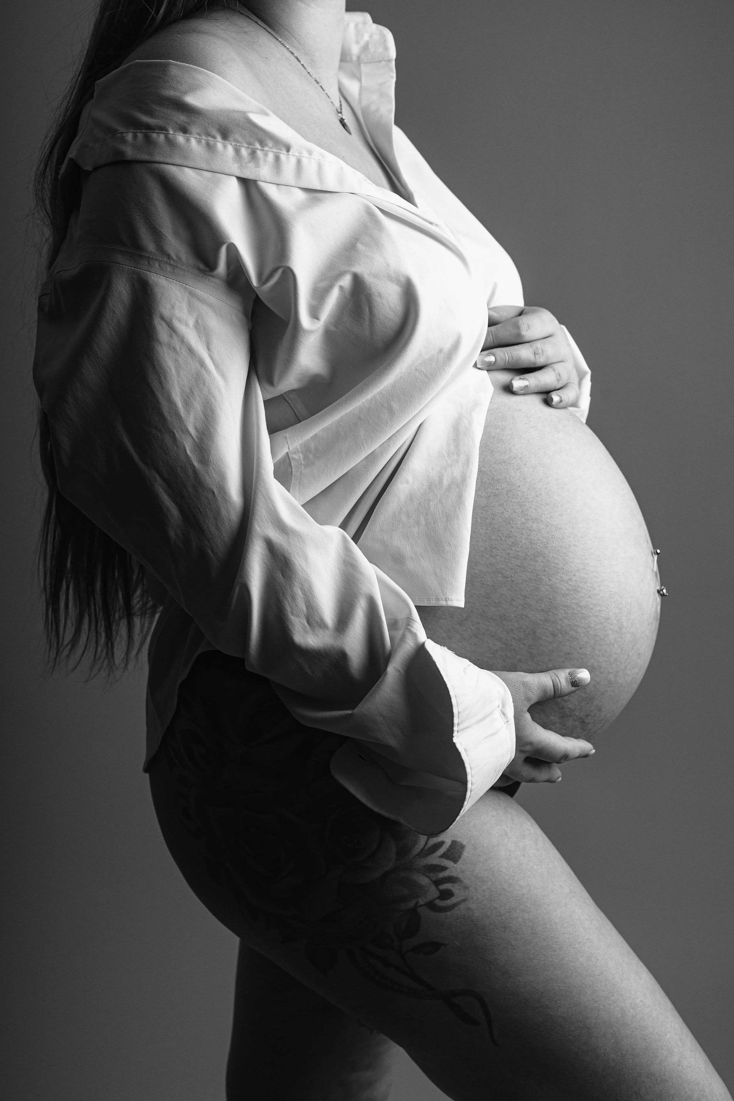 Black and white photo of a pregnant woman in a satin shirt, holding her belly with both hands, showcasing her tattooed arm and pregnant belly.
