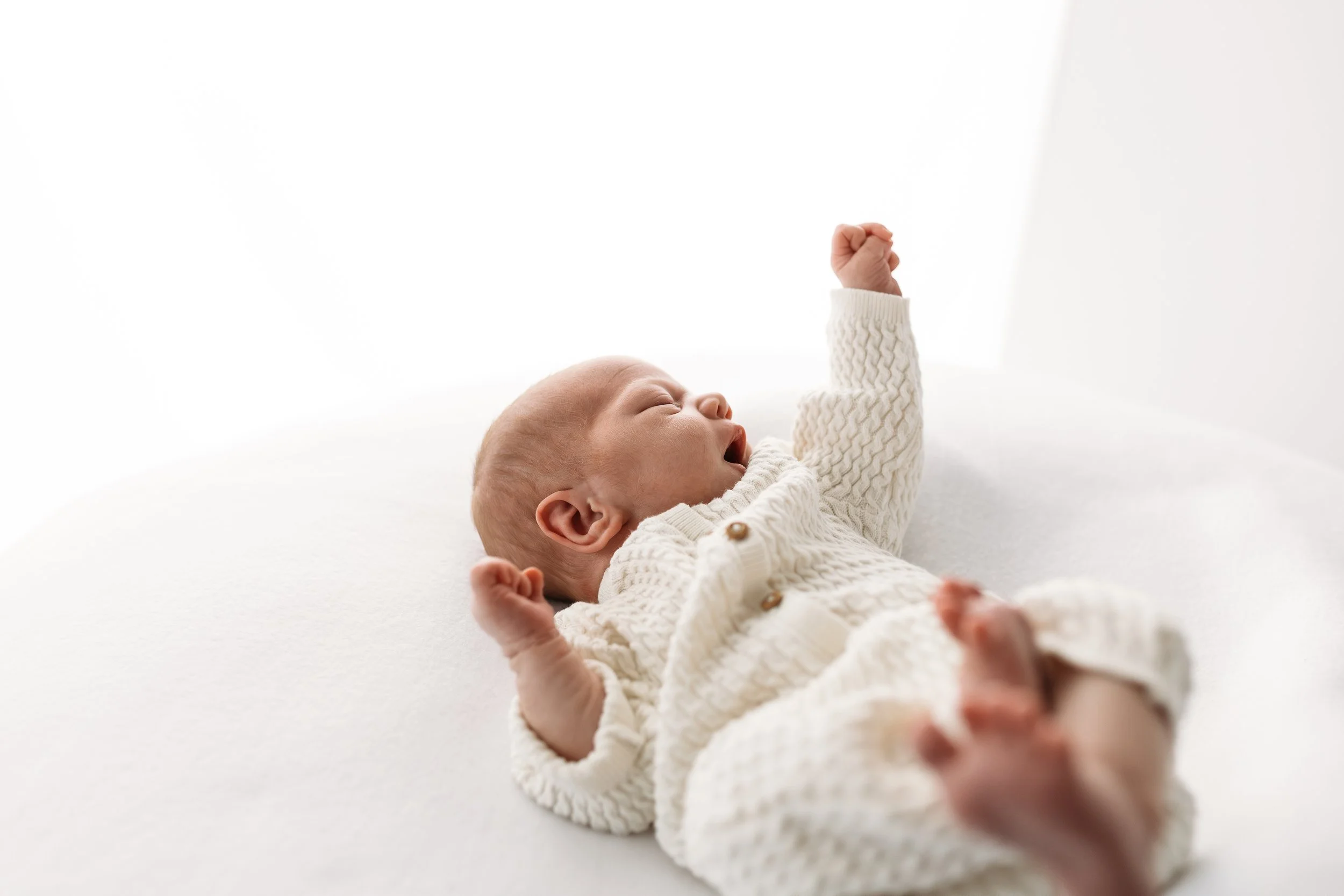 A baby lying on its back on a white surface, wearing a cream-colored knitted sweater, with one arm raised and mouth open.