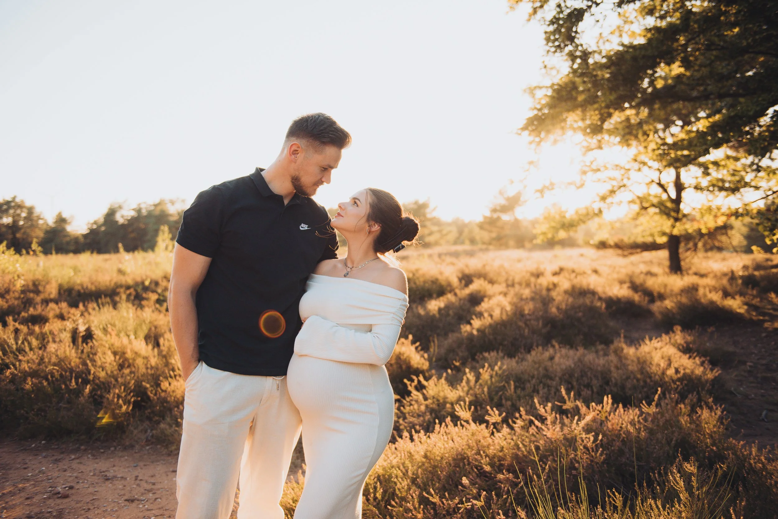 A couple stands close together in an outdoor field during sunset, looking into each other's eyes. The man wears a black Nike shirt and white pants, while the woman is dressed in a white off-the-shoulder top and white pants. The background features trees and golden sunlight.