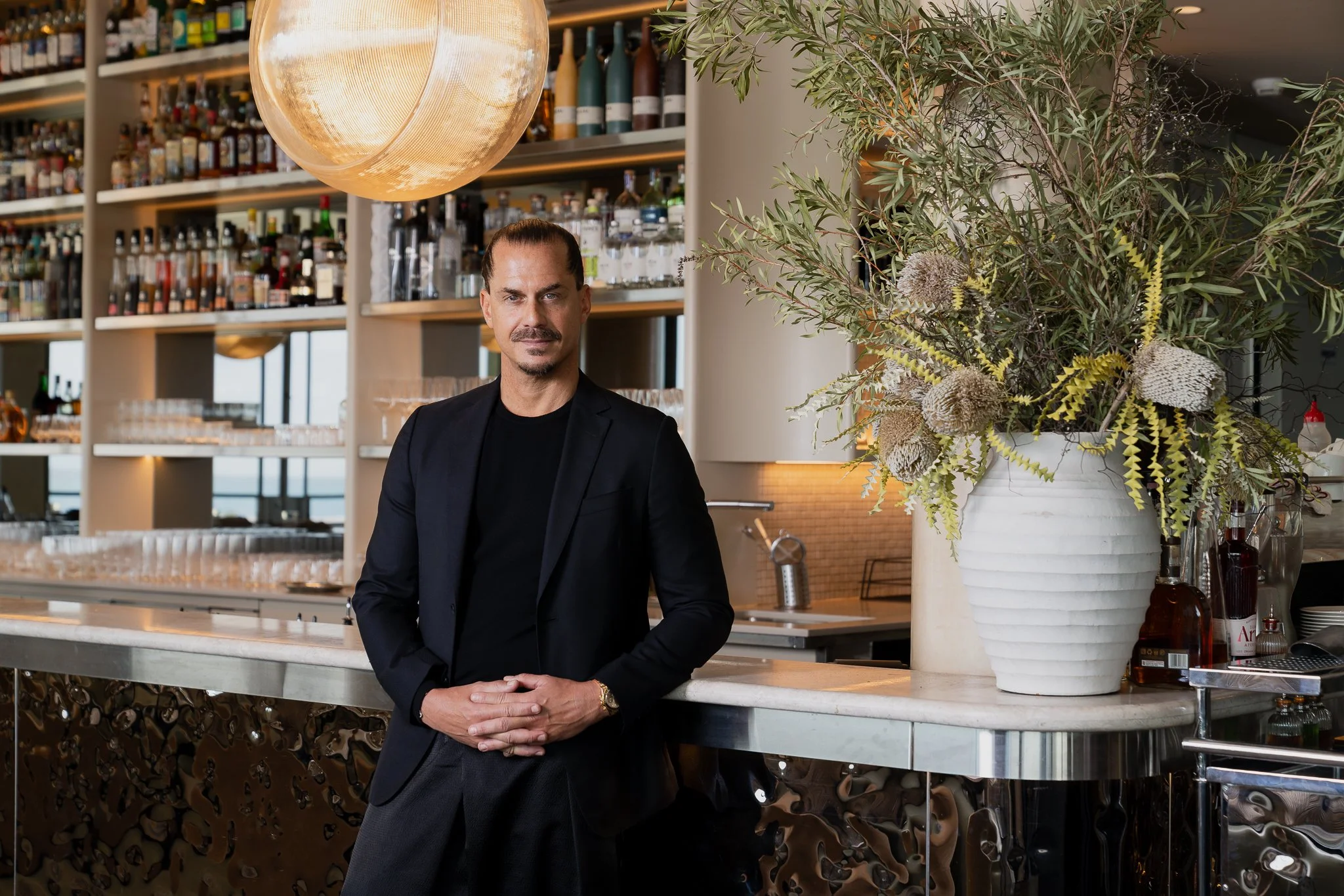 A man with slicked-back hair and a mustache, dressed in a black blazer and shirt, standing at a bar with a large white flower arrangement in a white pot, in a setting with shelves of liquor bottles in the background and warm lighting.