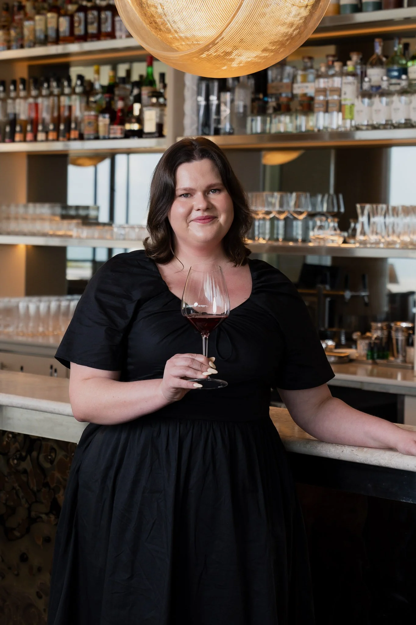 A woman with shoulder-length dark hair wearing a black dress, holding a glass of red wine, standing in a modern bar or restaurant setting with shelves of bottles and glassware in the background.