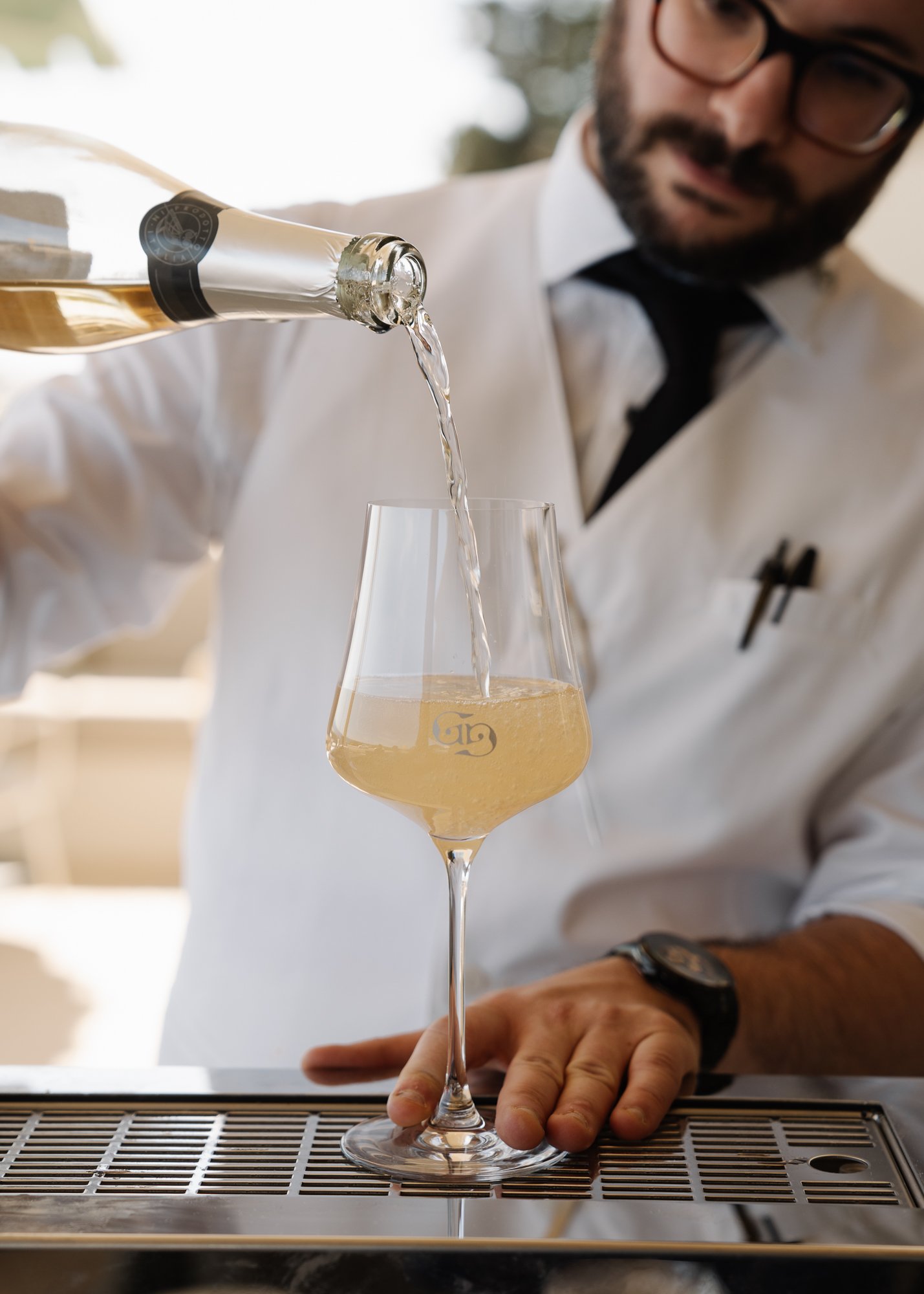 Bartender in white shirt and black tie pours a glass of sparkling white wine or champagne at a bar.