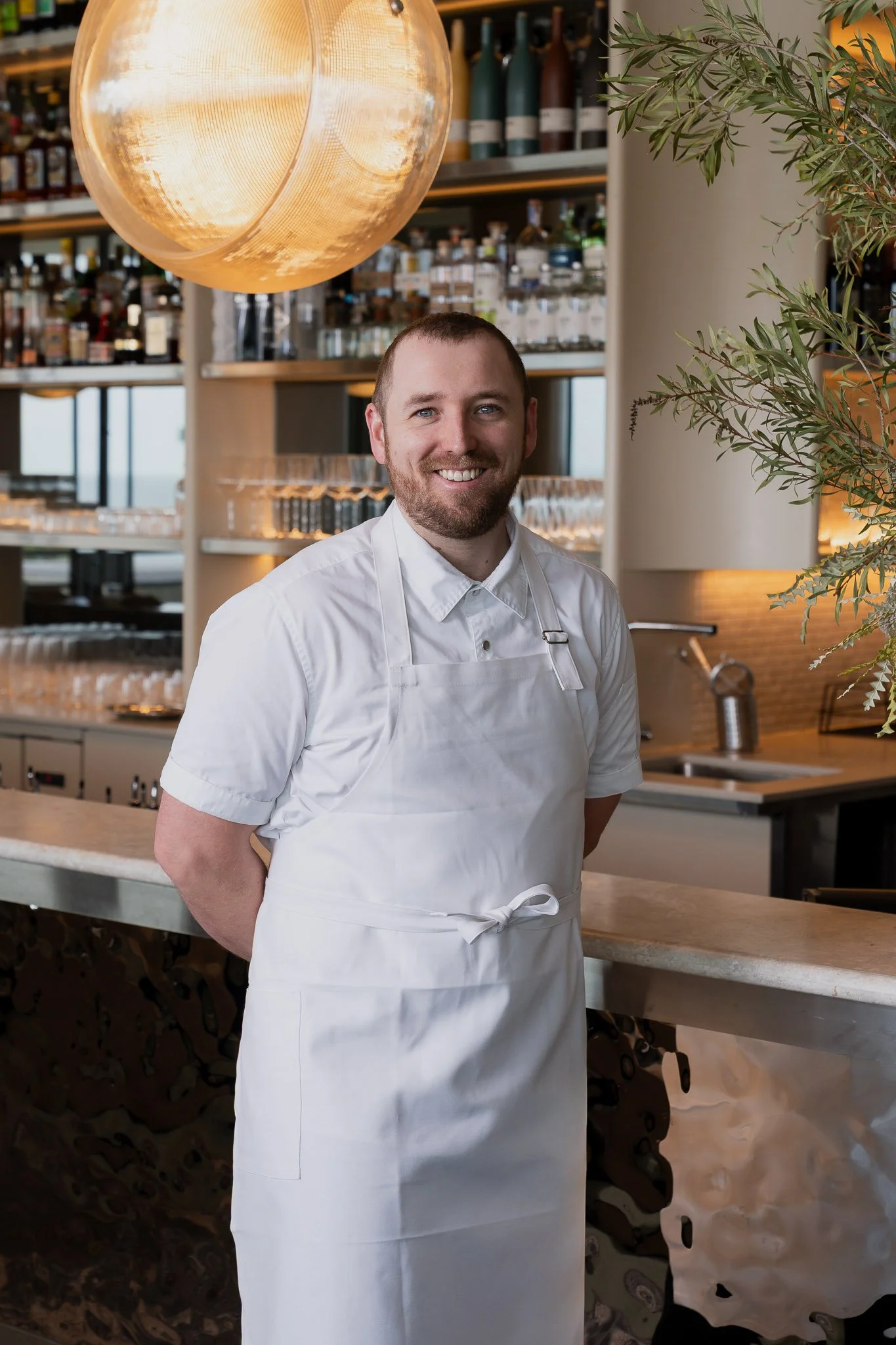 A smiling man in a white chef's uniform standing in a restaurant with shelves of bottles and glasses behind him, illuminated by a large round amber light fixture, with a plant on the right side.