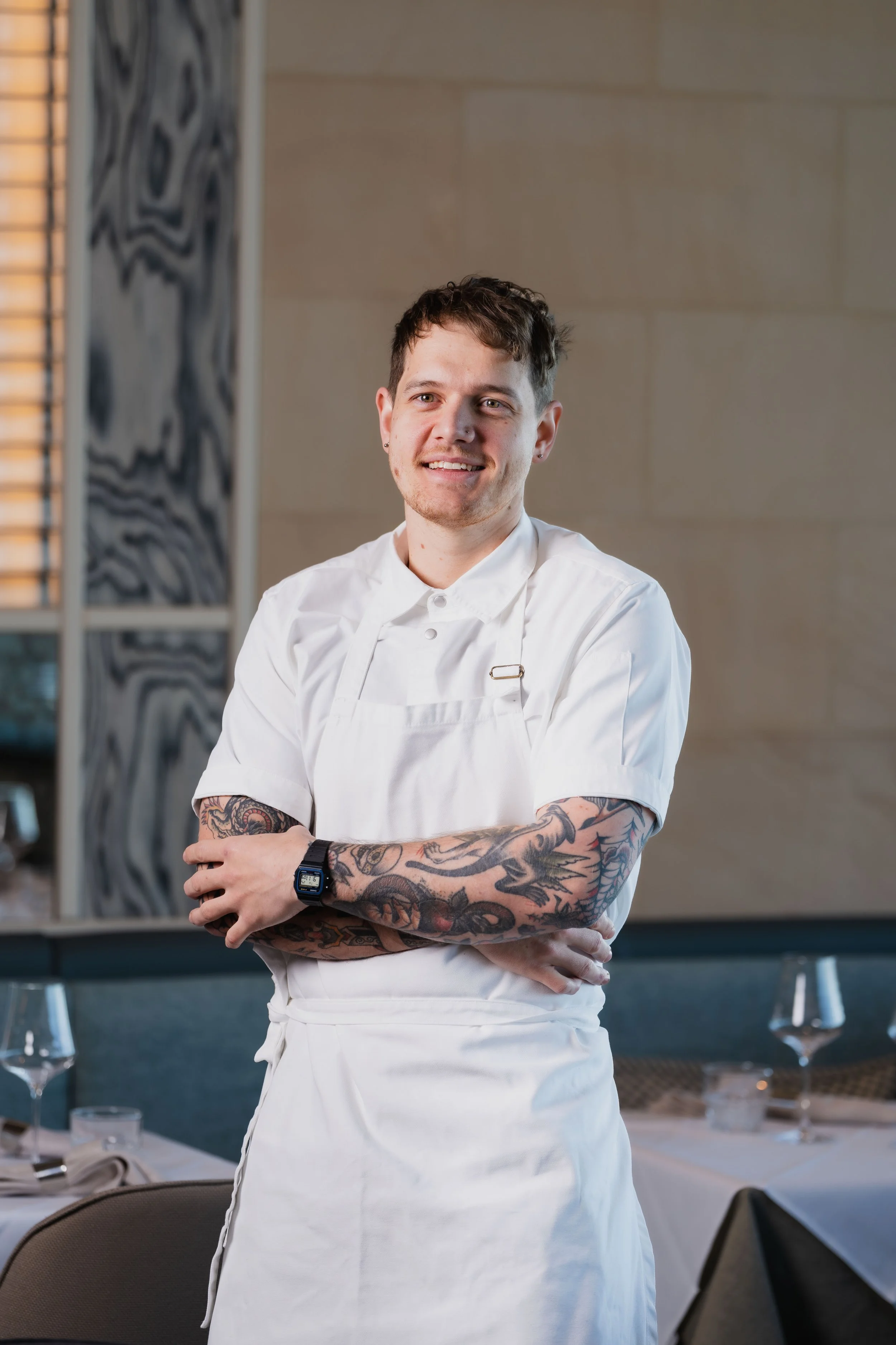 Young male chef with tattoos on arms, wearing a white chef uniform and apron, standing with arms crossed in a restaurant setting with tables, glasses, and chairs.