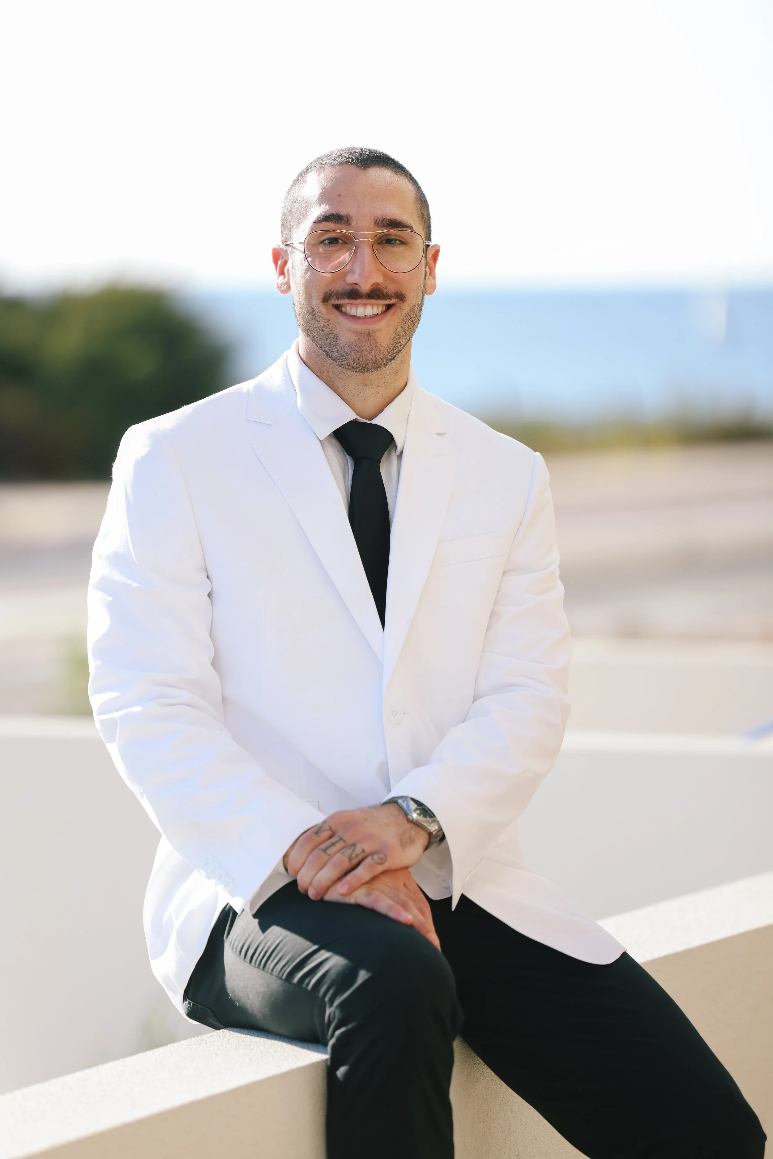 Smiling man wearing glasses, white suit jacket, black tie, black pants, sitting outdoors on a ledge with a blurred background of greenery and blue sky.