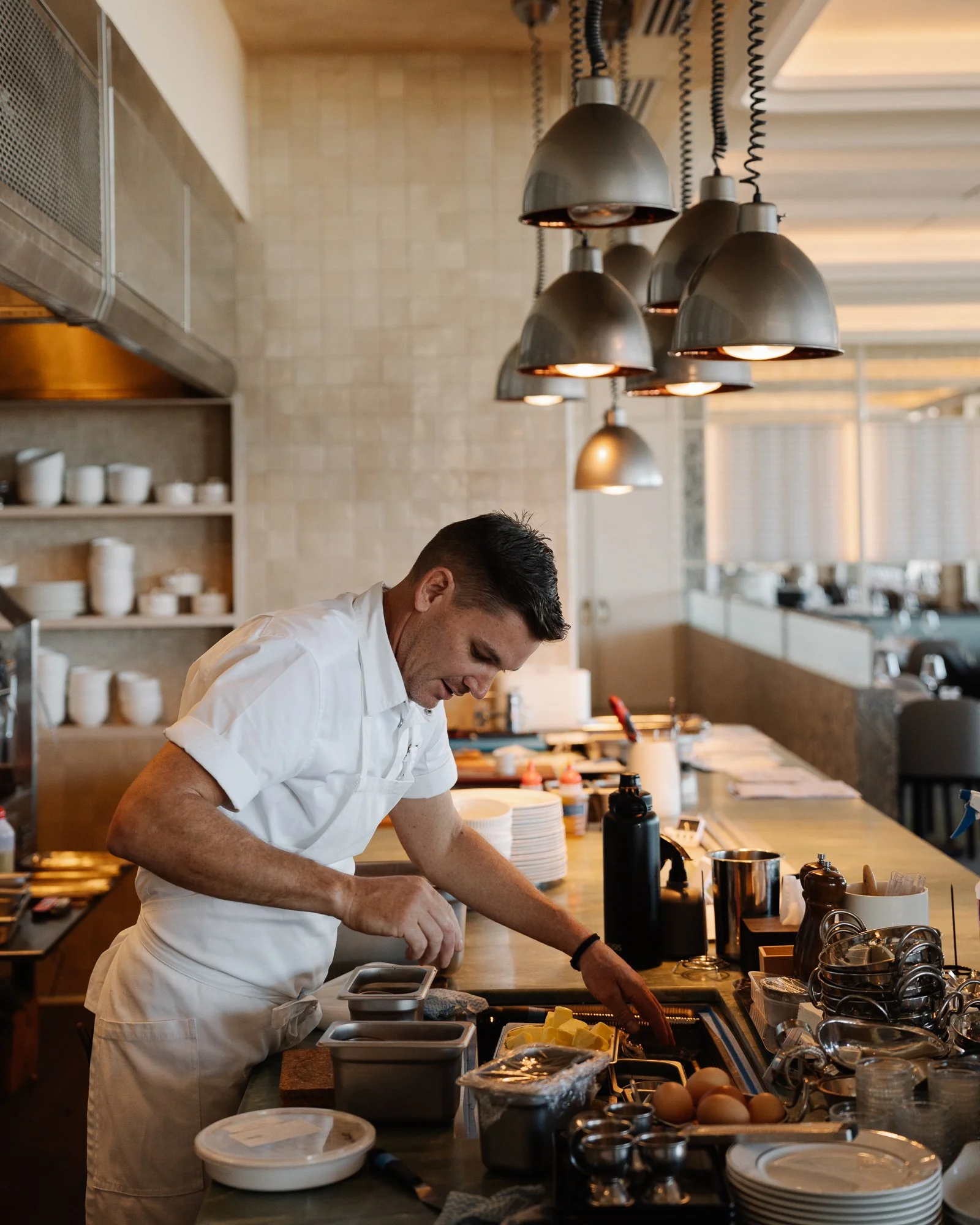 A chef in a white uniform is working in a professional kitchen, preparing ingredients at a counter with various cooking utensils and supplies.