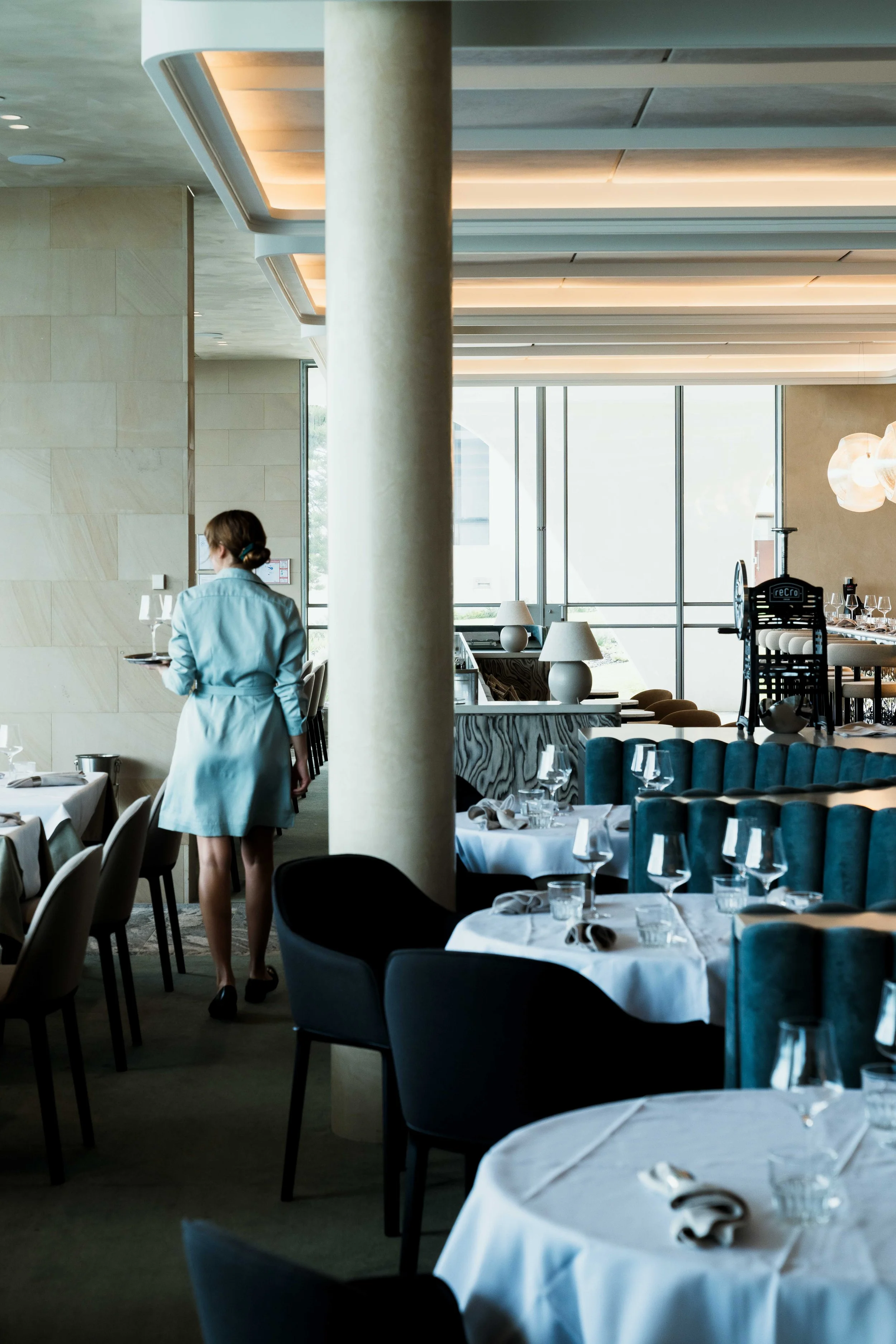 Interior of a restaurant with tables set for dining, featuring white tablecloths, wine glasses, and neatly folded napkins. A woman in a light blue dress, with her hair tied back, is walking away from the camera carrying a tray. The decor includes large windows, modern lighting fixtures, and a combination of chairs and sofas.