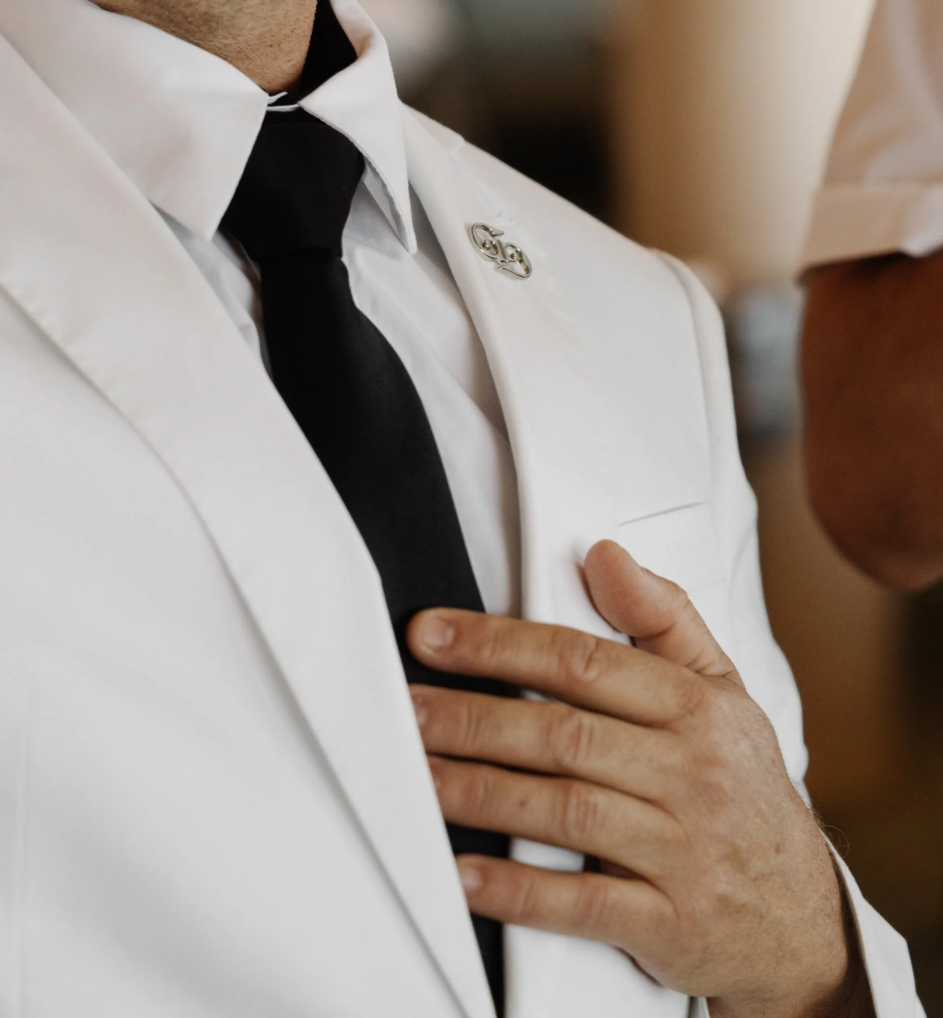 A close-up of a person's hand touching the white lapel of a white shirt, which is accessorized with a decorative silver pin, with a black tie visible underneath.