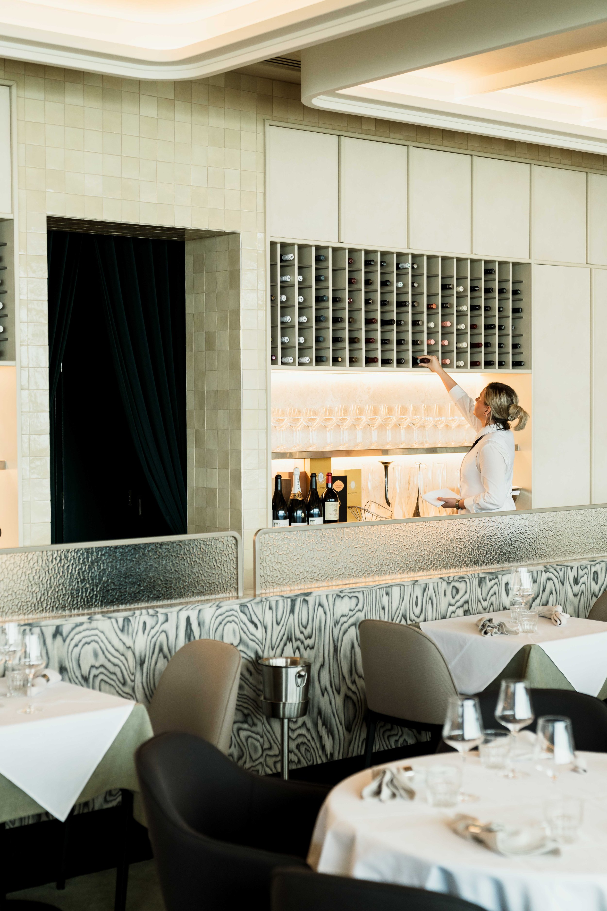 Interior of a fine dining restaurant with tables set for guests, some with white tablecloths, napkins, and glasses. A woman with blonde hair, dressed in white, is reaching for a wine bottle from a wall-mounted wine rack with multiple bottles. The room has modern decor with a marble-patterned panel and frosted glass divider.