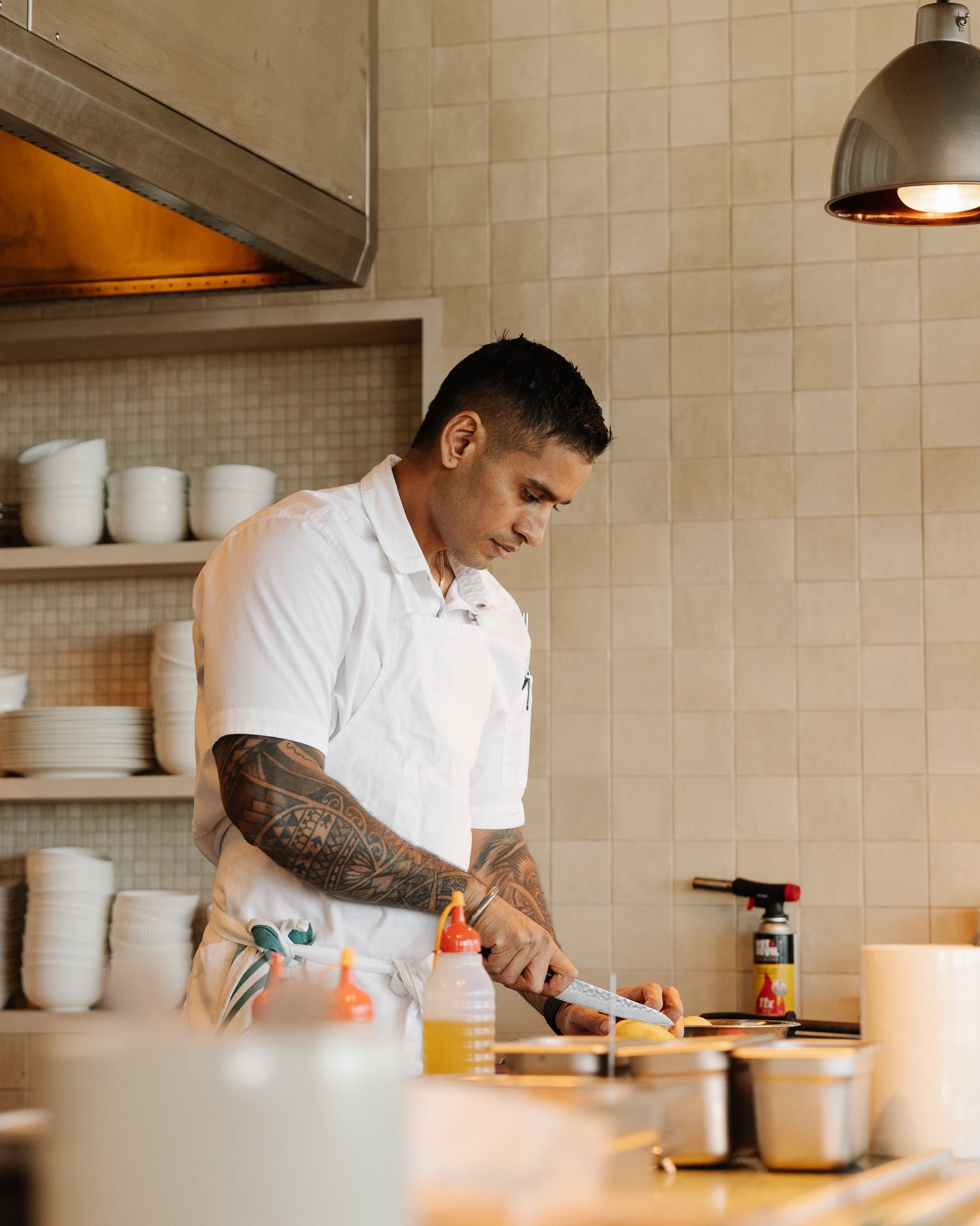 A man with tattoos on his arms preparing food in a kitchen, wearing a white shirt and apron, with shelves of bowls and condiments around him.