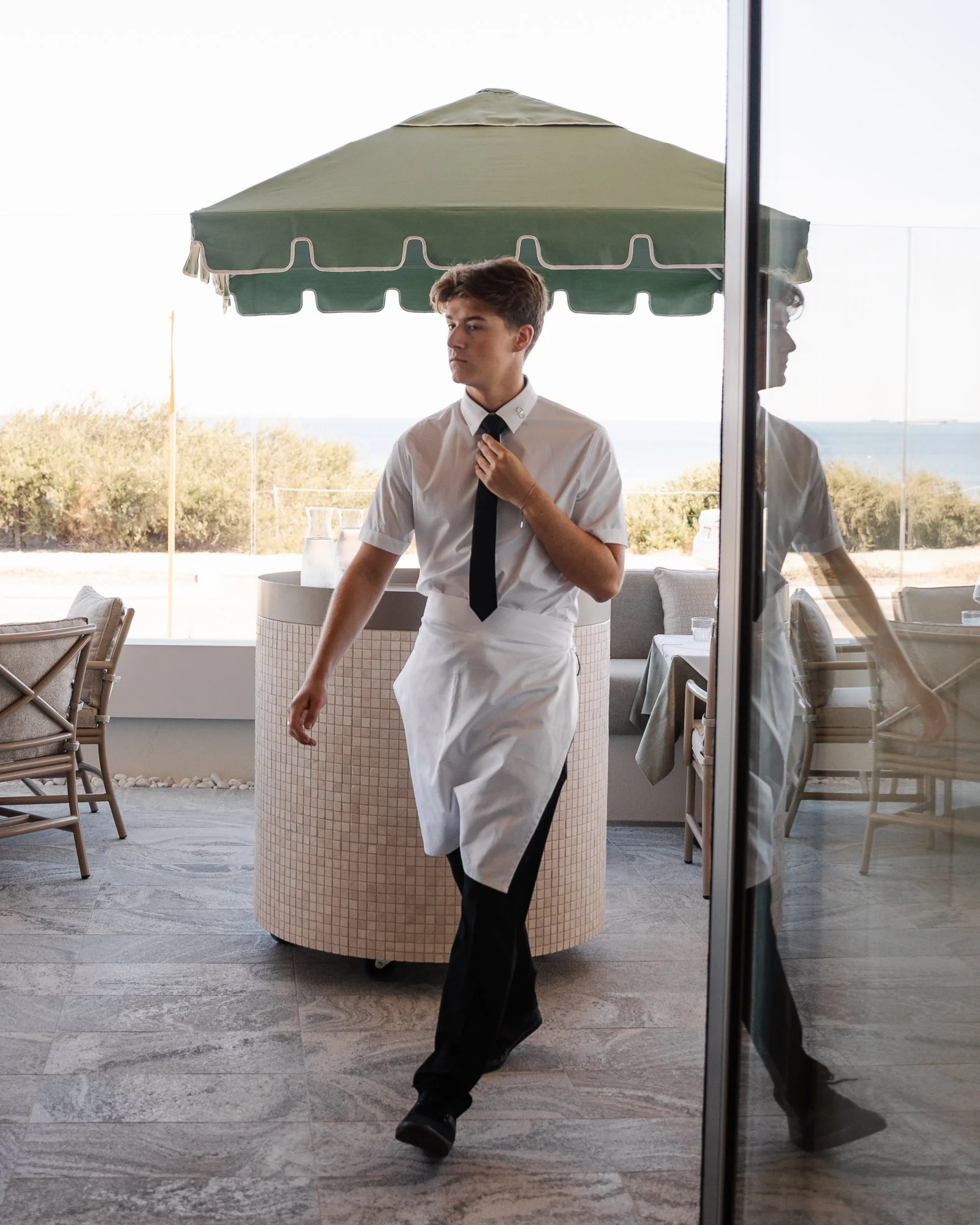 A young man wearing a white shirt, black tie, and apron, standing inside a restaurant or cafe near a window and glass door, with outdoor seating and an umbrella visible outside.