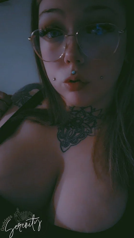 A woman with glasses and tattoos taking a selfie, with dark hair and makeup, in a dimly lit room.