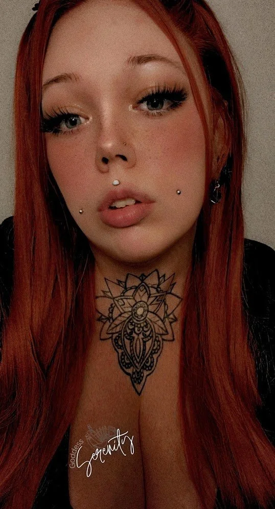 Close-up of a woman with long red hair, blue eyes, pierced nose, and jewelry, wearing makeup, with a tattoo on her chest, and a dark top.