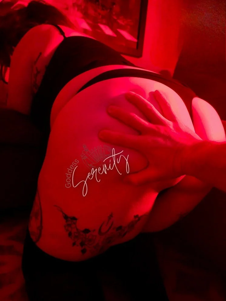 A person with tattoos on their side and back, wearing black underwear, lying on their side in a red-lit room, with their hand on their buttocks, and a tattoo reading 'goodness' and 'serenity' visible on their side.